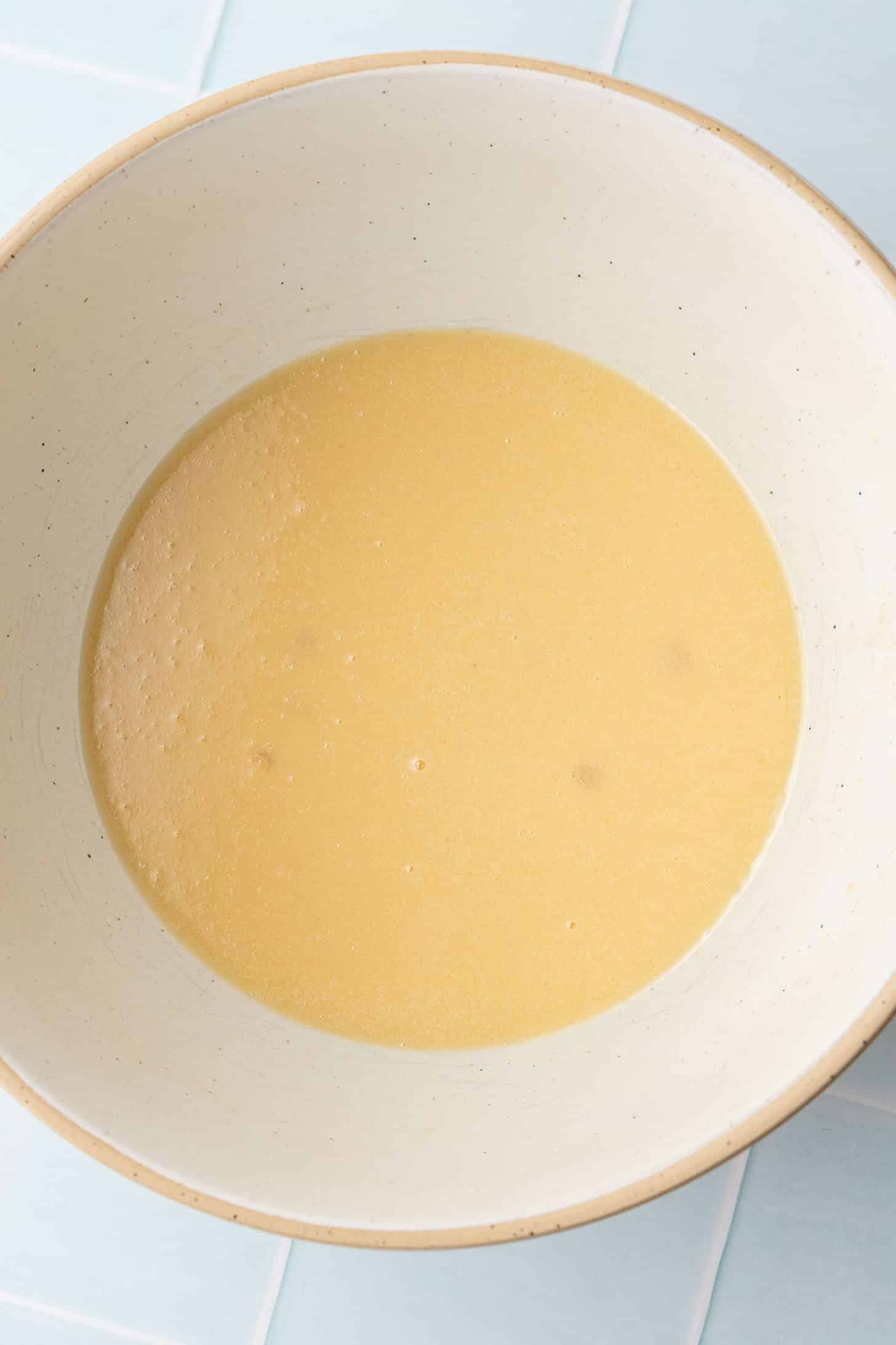 Wet muffin ingredients including melted butter, eggs, brown sugar, and milk whisked together until smooth and golden in a cream-colored ceramic bowl