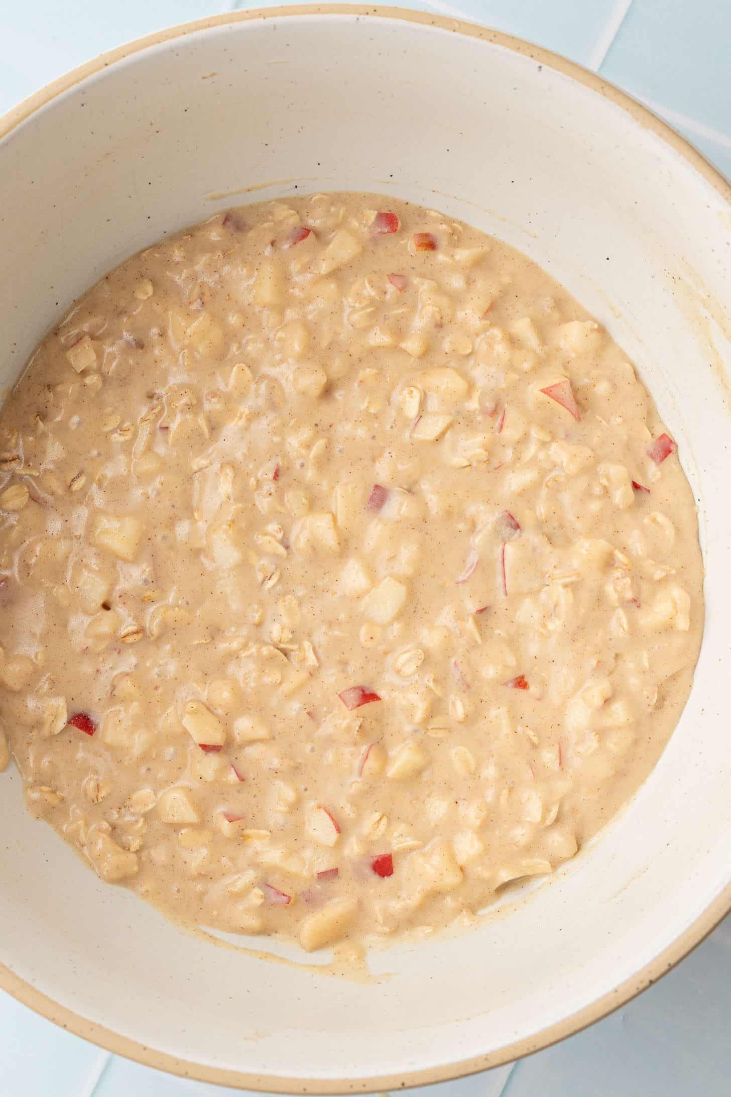 Completed apple oatmeal muffin batter with rolled oats and diced apple pieces folded throughout in a cream-colored ceramic bowl