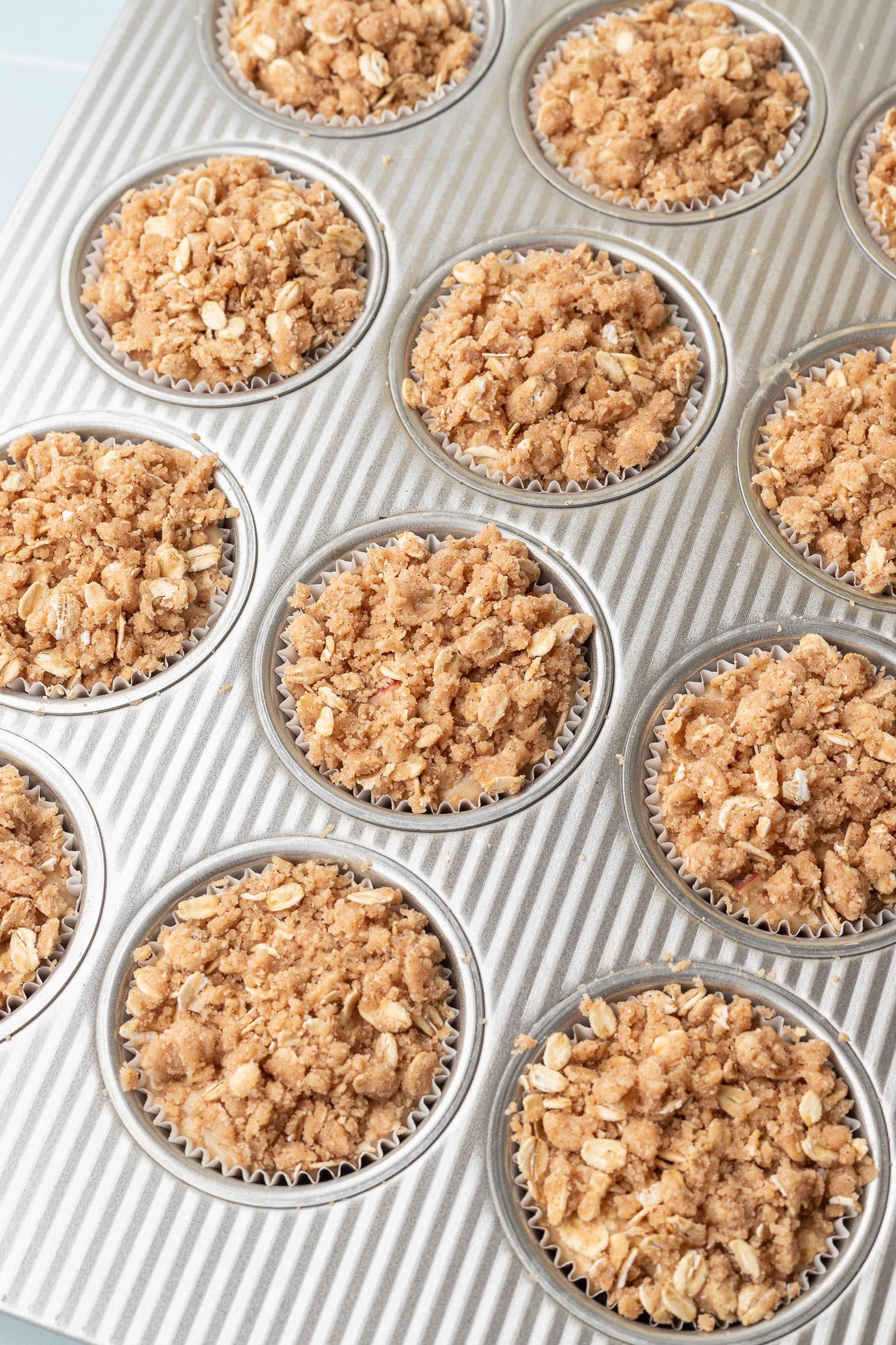 Apple oat muffin batter topped with a generous layer of cinnamon oat streusel in a silver muffin tin lined with white parchment liners, ready to bake
