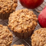 Freshly baked apple oat muffins with golden cinnamon streusel tops cooling on a wire rack, with whole red apples in the background