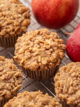 Freshly baked apple oat muffins with golden cinnamon streusel tops cooling on a wire rack, with whole red apples in the background