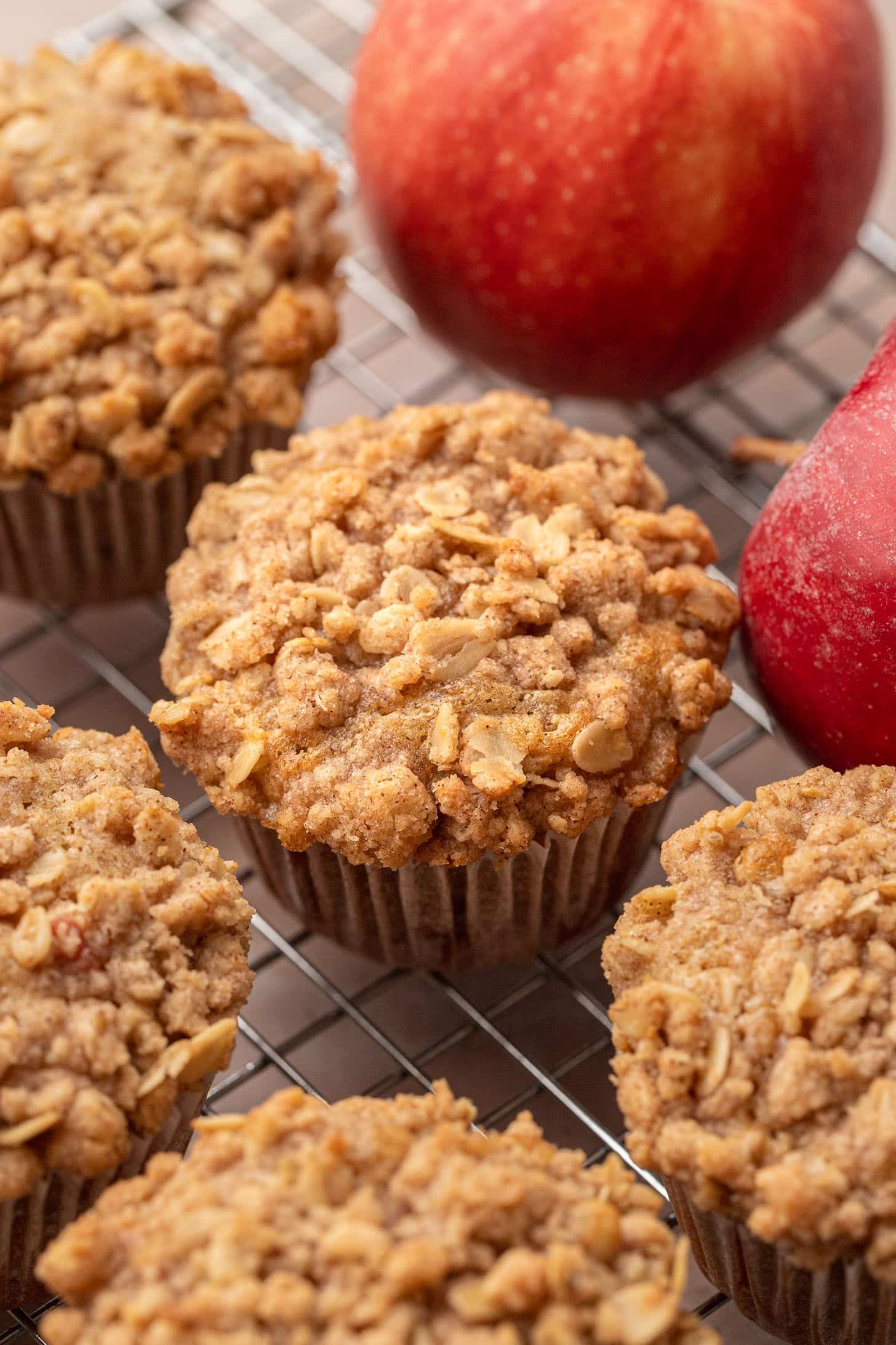 Freshly baked apple oat muffins with golden cinnamon streusel tops cooling on a wire rack, with whole red apples in the background