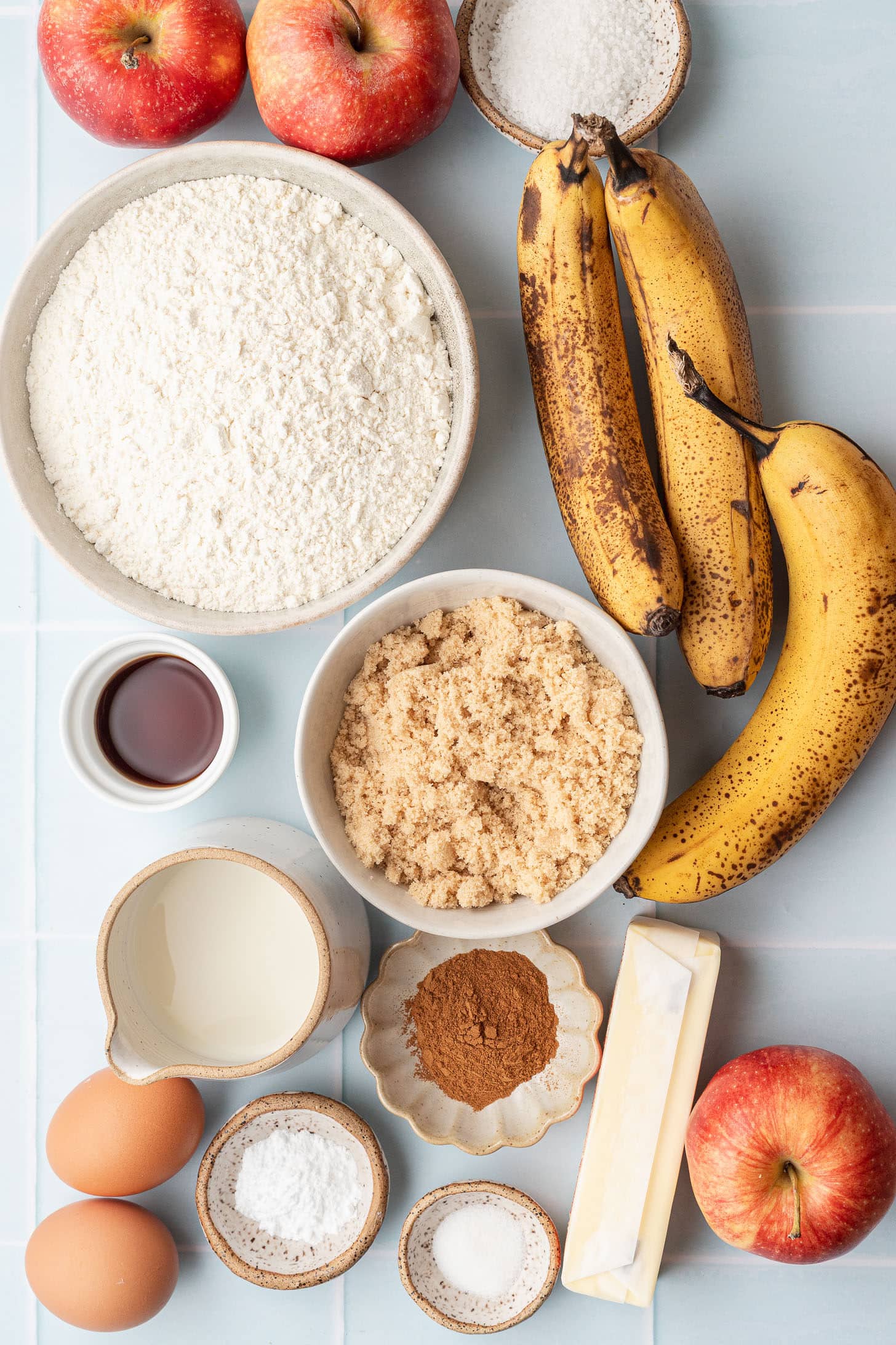 Flat lay of apple banana muffin ingredients including red apples, overripe bananas, flour, brown sugar, butter, eggs, milk, vanilla extract, cinnamon, baking powder, and salt on a light blue tile surface
