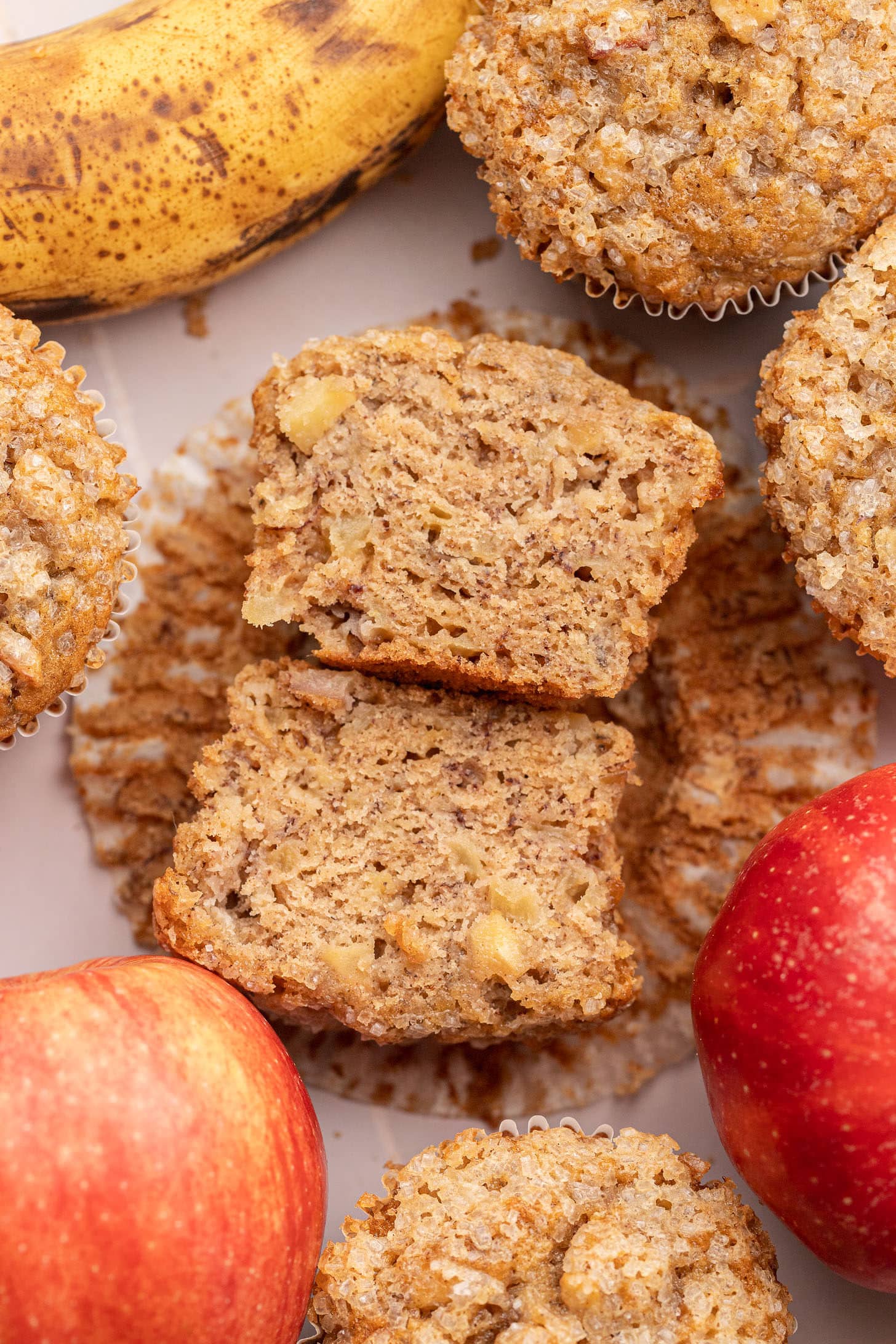 Two apple banana muffins stacked and split open to show the soft, moist crumb with apple pieces throughout and a sparkling sugar crust, surrounded by more muffins and fresh red apples