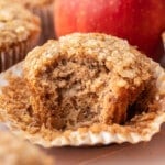 Close-up of an apple banana muffin with its wrapper peeled back, broken open to reveal a tender crumb packed with diced apple pieces and finished with a crunchy coarse sugar top, with a red apple in the background