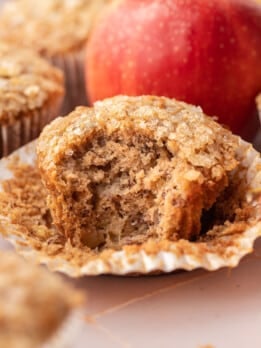 Close-up of an apple banana muffin with its wrapper peeled back, broken open to reveal a tender crumb packed with diced apple pieces and finished with a crunchy coarse sugar top, with a red apple in the background