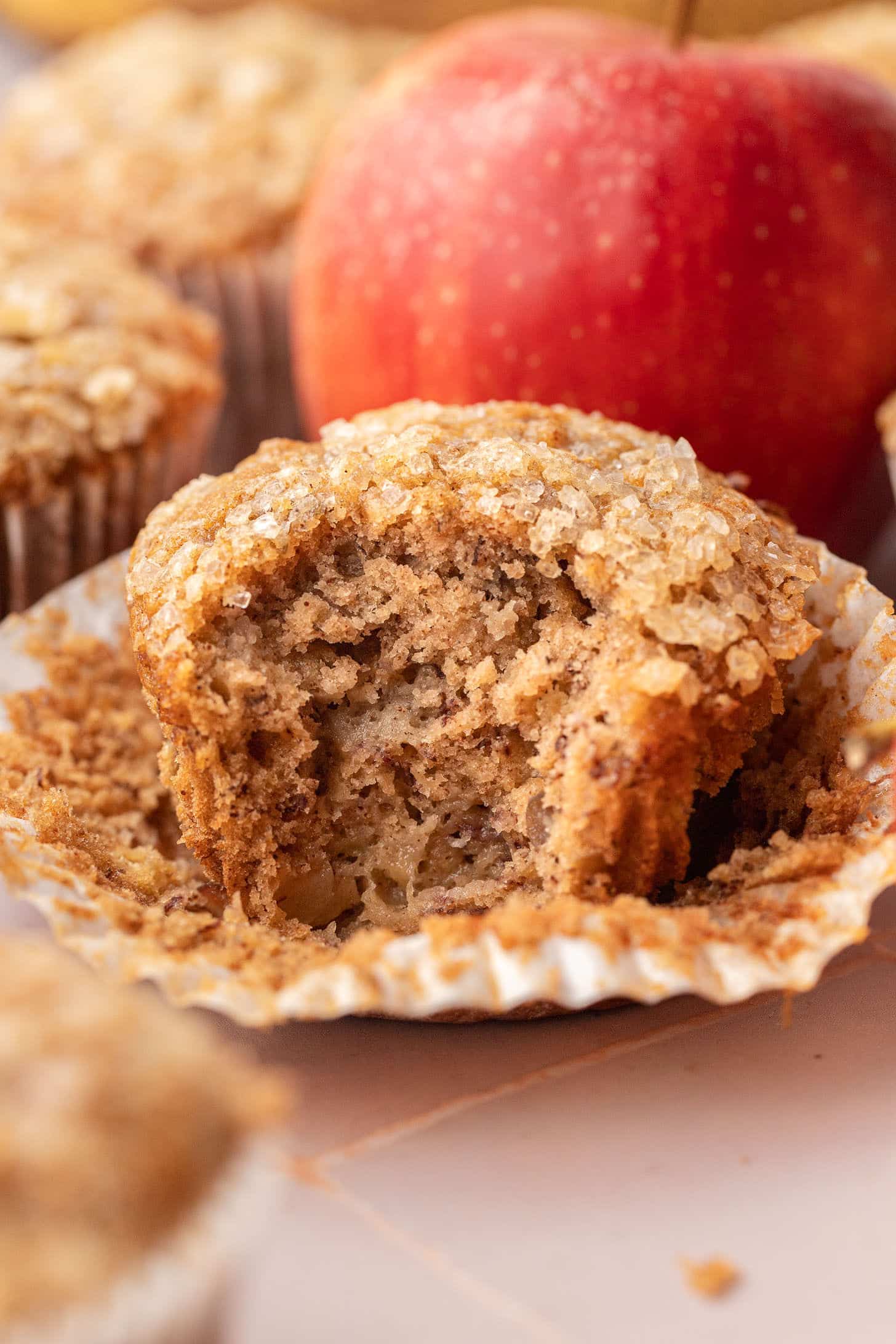 Close-up of an apple banana muffin with its wrapper peeled back, broken open to reveal a tender crumb packed with diced apple pieces and finished with a crunchy coarse sugar top, with a red apple in the background