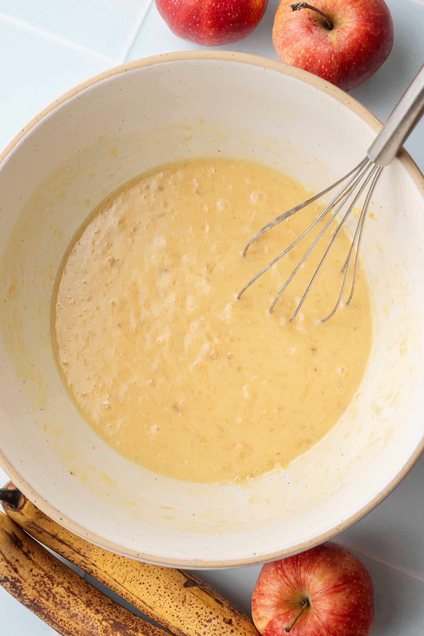 Wet muffin ingredients including mashed banana, butter, eggs, brown sugar, and milk whisked together until smooth in a cream-colored ceramic bowl, with red apples and overripe bananas nearby