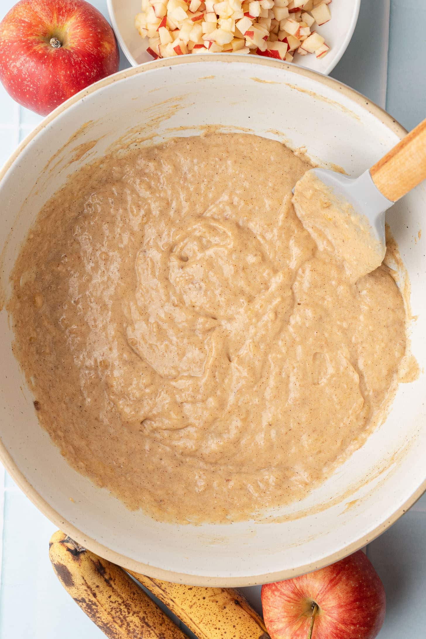 Apple banana muffin batter with dry ingredients folded in, thick and smooth with a spatula resting in the bowl, with a bowl of diced apples and overripe bananas alongside