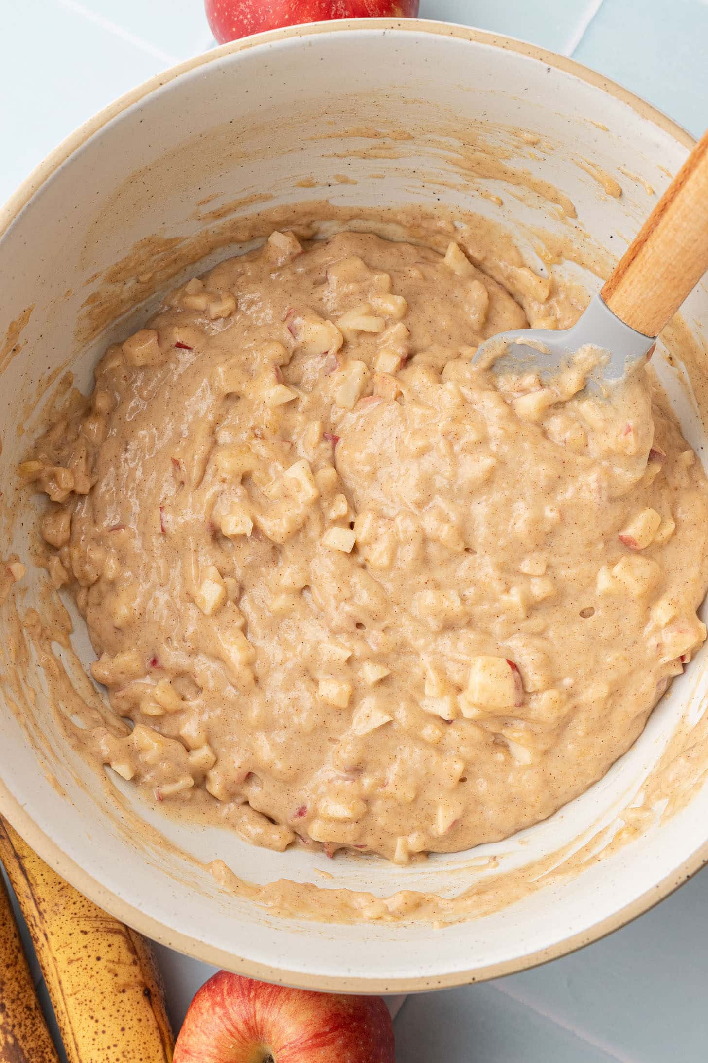 Completed apple banana muffin batter with diced apple pieces folded throughout in a cream-colored ceramic bowl, with a spatula, overripe bananas, and a red apple nearby