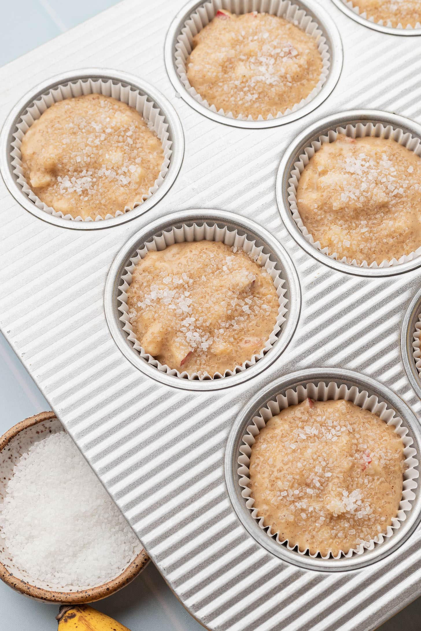 Apple banana muffin batter divided into a silver muffin tin lined with white parchment liners and sprinkled with coarse turbinado sugar on top, with a small bowl of sugar and a banana nearby