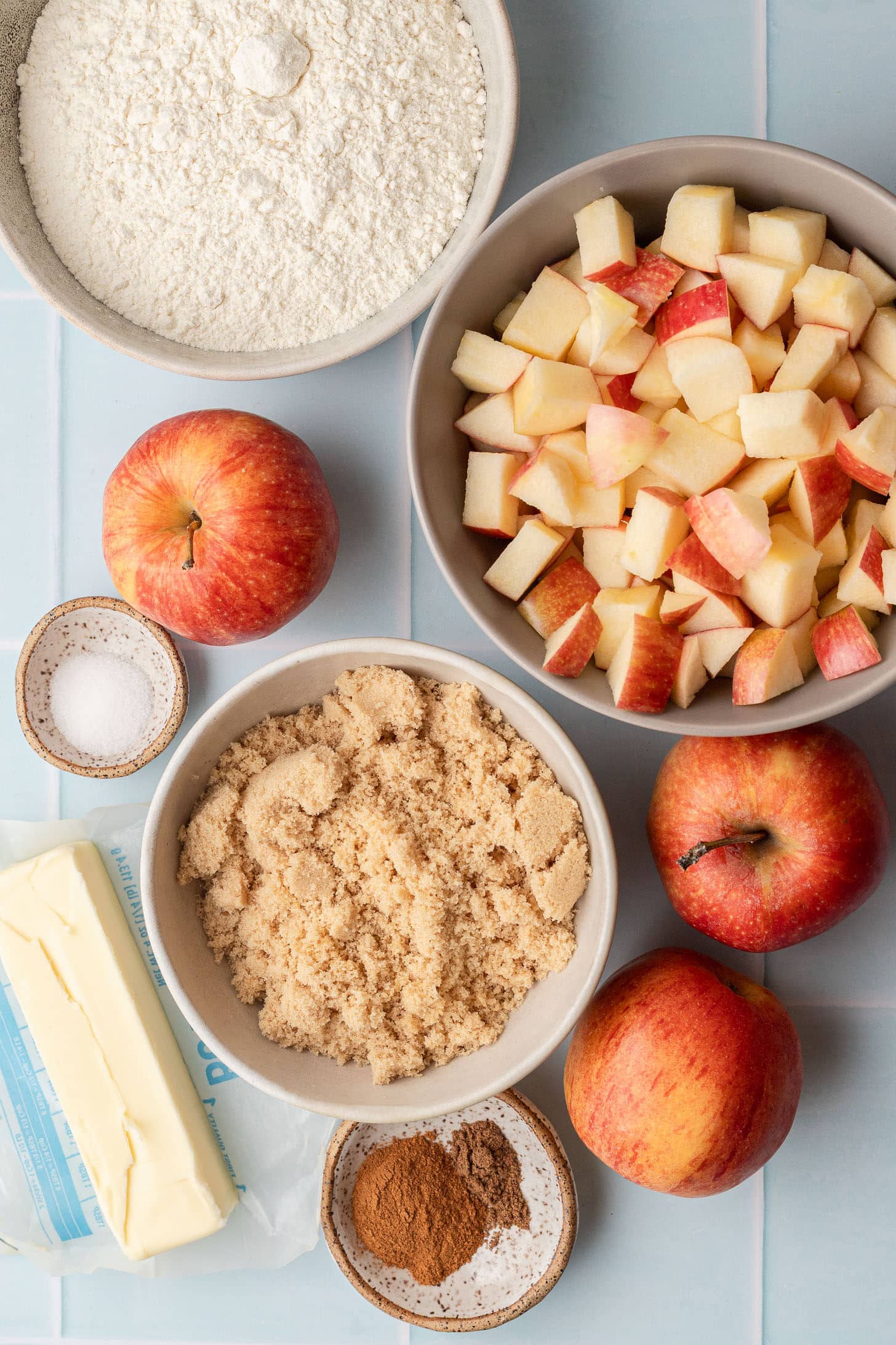 Flat lay of apple crumble ingredients including diced apples in a bowl, a whole red apple, flour, brown sugar, a stick of butter, salt, and cinnamon on a light blue tile surface