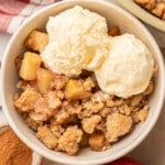 Overhead view of a serving of apple crumble in a white ceramic bowl topped with two generous scoops of vanilla ice cream, surrounded by fresh apples and a small dish of cinnamon