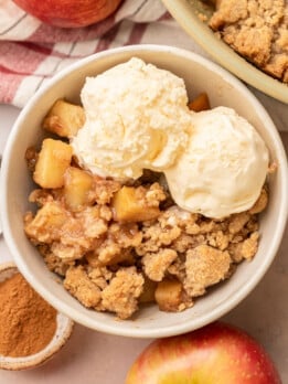 Overhead view of a serving of apple crumble in a white ceramic bowl topped with two generous scoops of vanilla ice cream, surrounded by fresh apples and a small dish of cinnamon