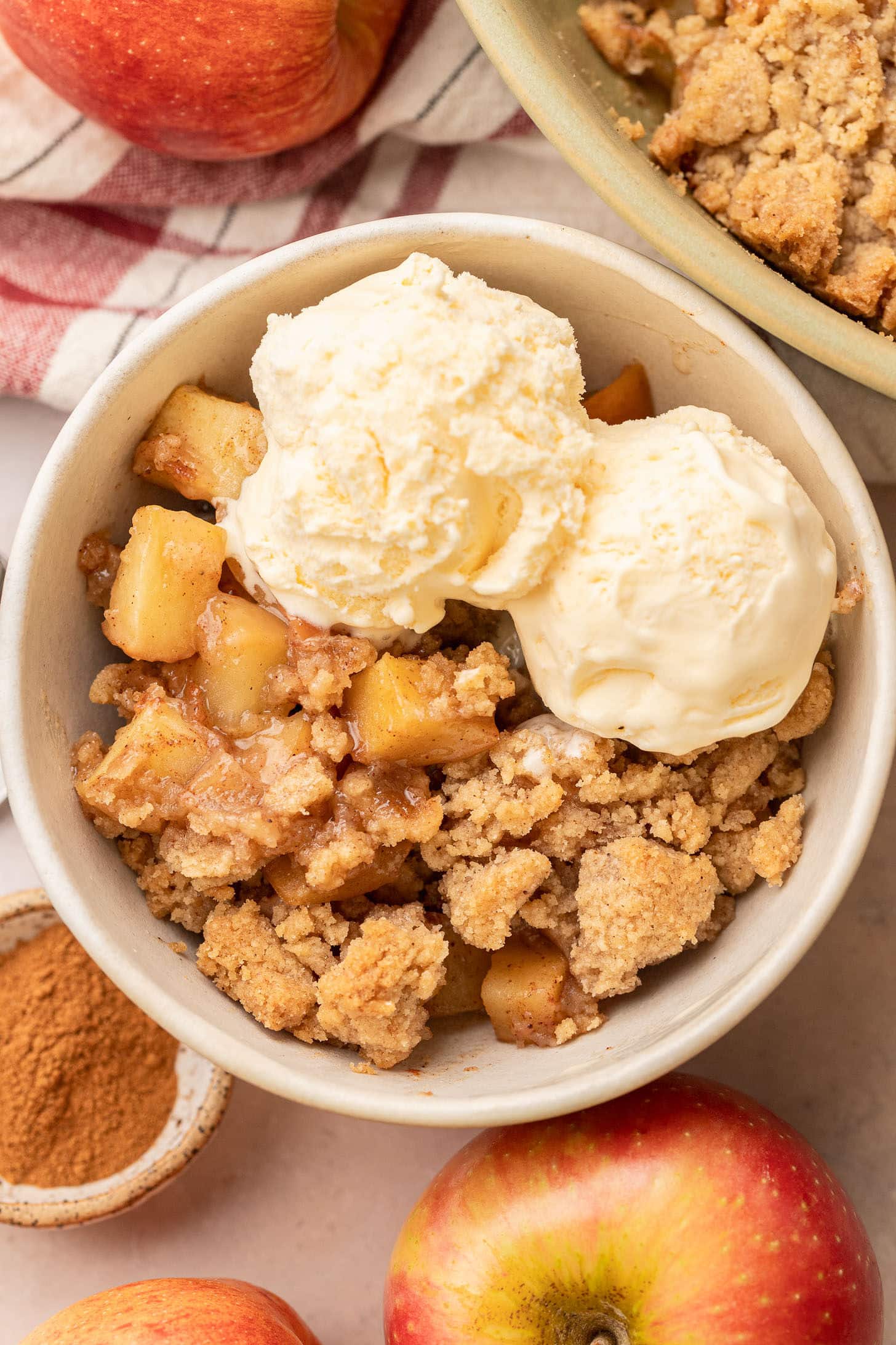 Overhead view of a serving of apple crumble in a white ceramic bowl topped with two generous scoops of vanilla ice cream, surrounded by fresh apples and a small dish of cinnamon
