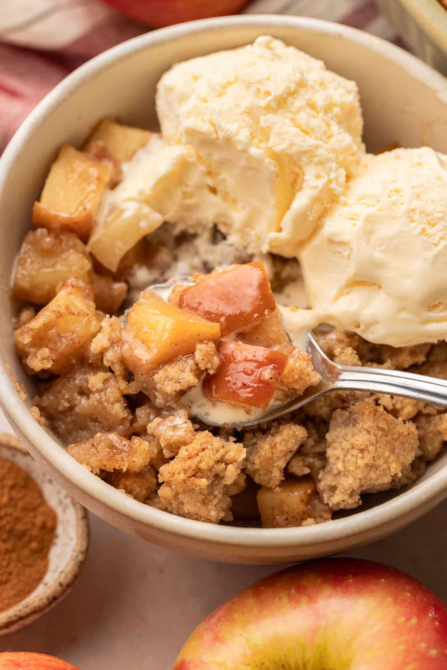 Close-up of a spoonful of apple crumble with juicy cinnamon apple pieces and crumble topping being lifted from a serving bowl topped with melting vanilla ice cream
