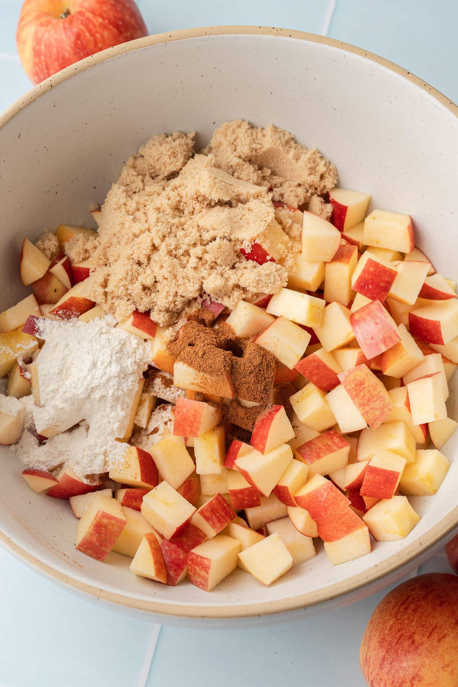 Diced red apples in a cream-colored bowl topped with brown sugar, flour, and cinnamon, before being tossed together for the filling
