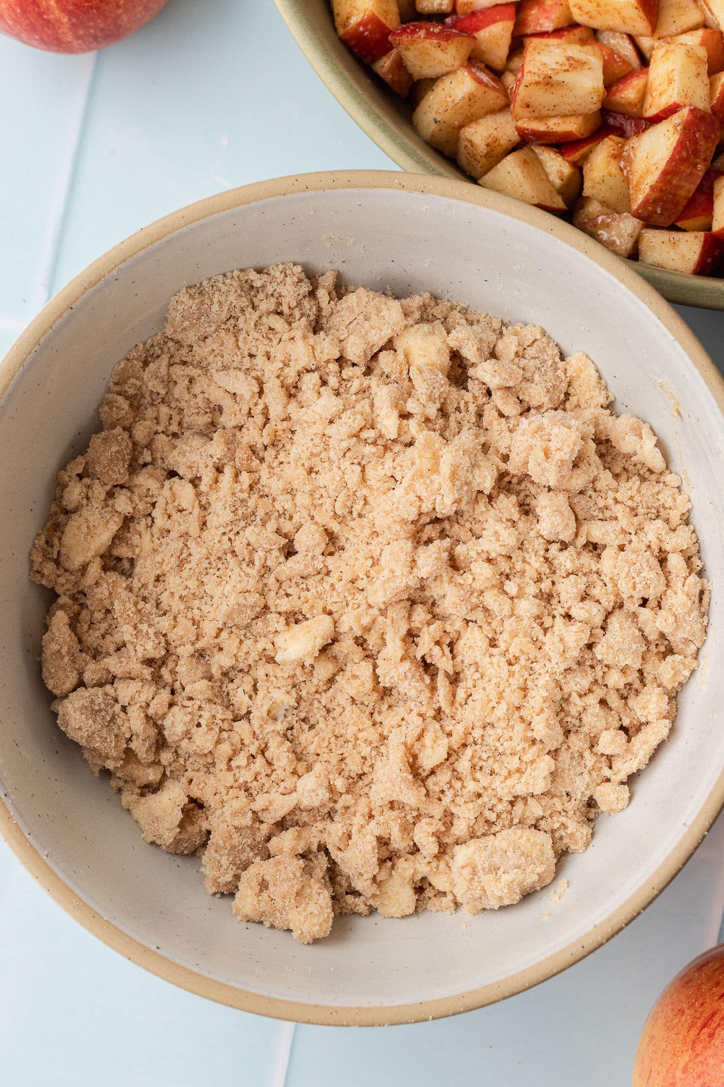 Sandy cinnamon brown sugar crumble topping mixed to a crumbly texture in a cream-colored speckled bowl, with the cinnamon apple filling visible in the background