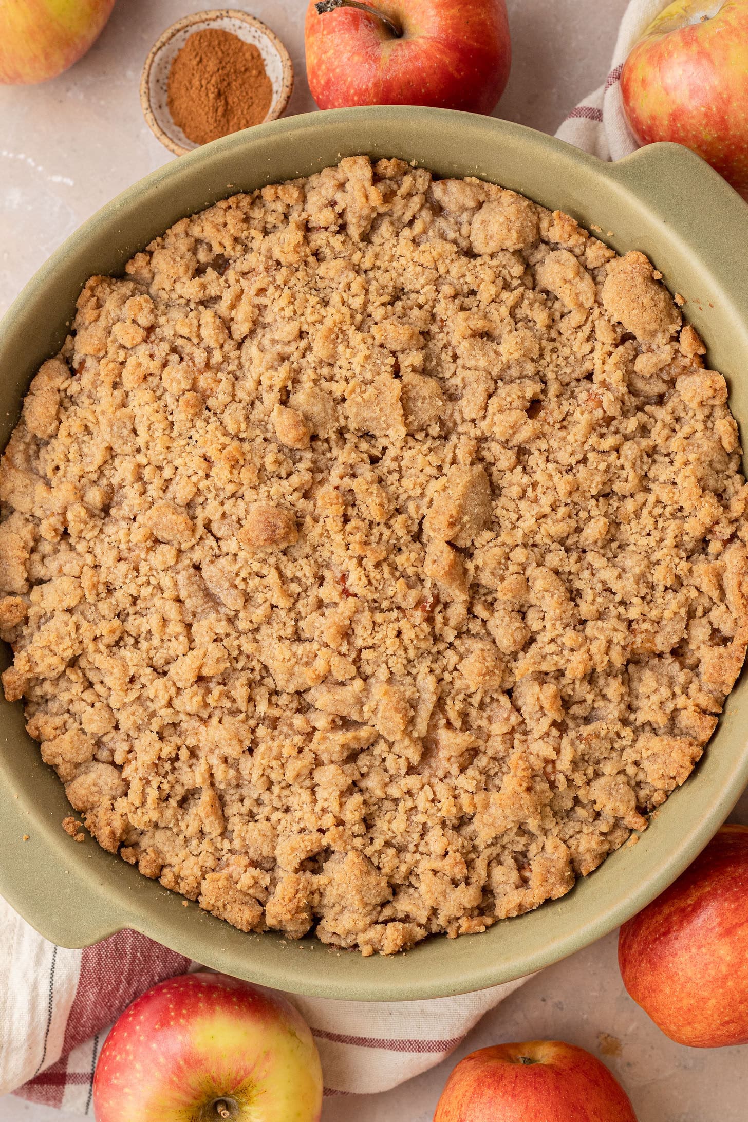 Freshly baked apple crumble with a golden cinnamon crumble topping in a green ceramic baking dish, surrounded by fresh red apples and a small dish of cinnamon