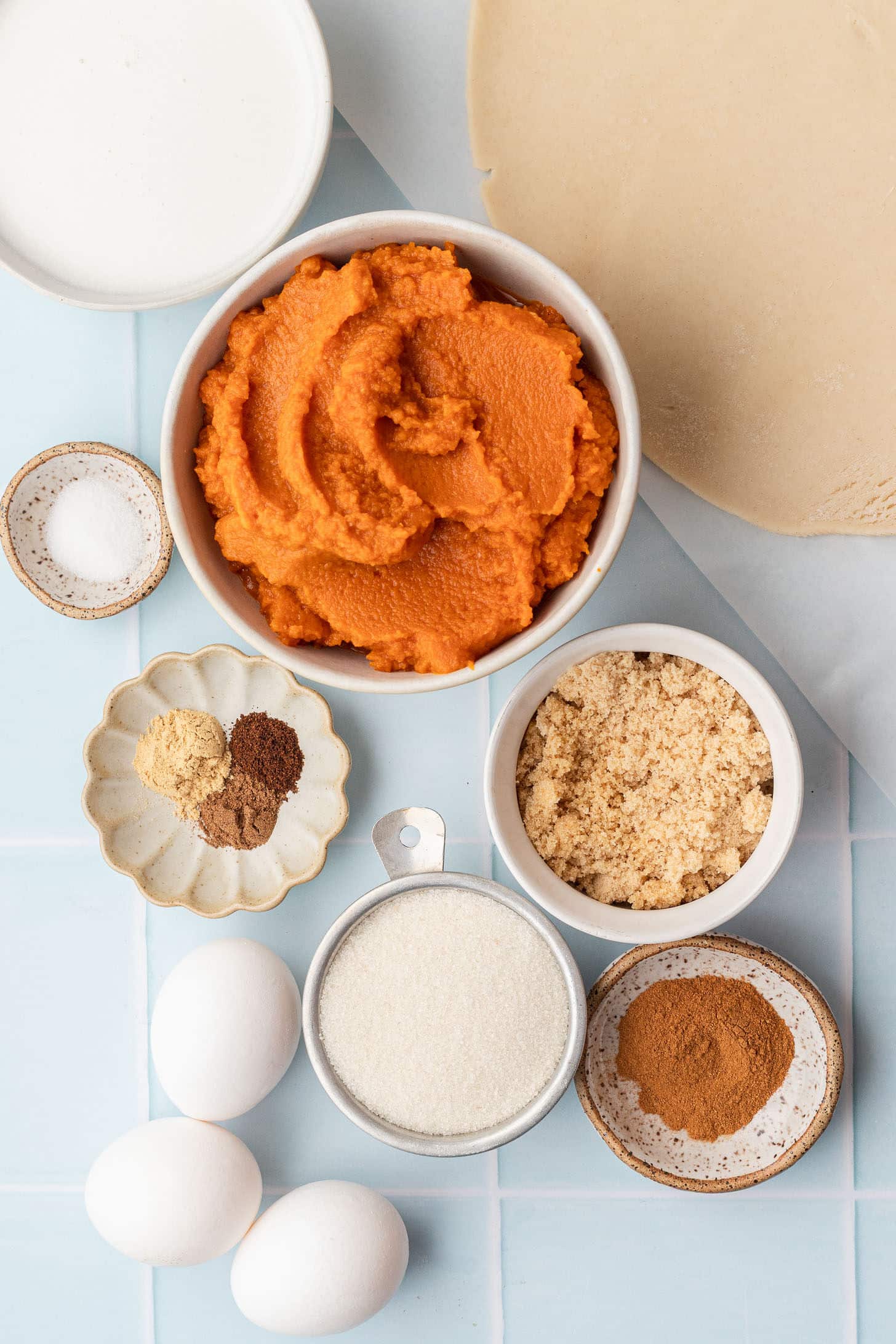 Flat lay of pumpkin pie ingredients including a can of pumpkin puree, can of coconut milk, eggs, brown sugar, white sugar, cinnamon, nutmeg, ginger, salt, and a store-bought pie crust on a light surface