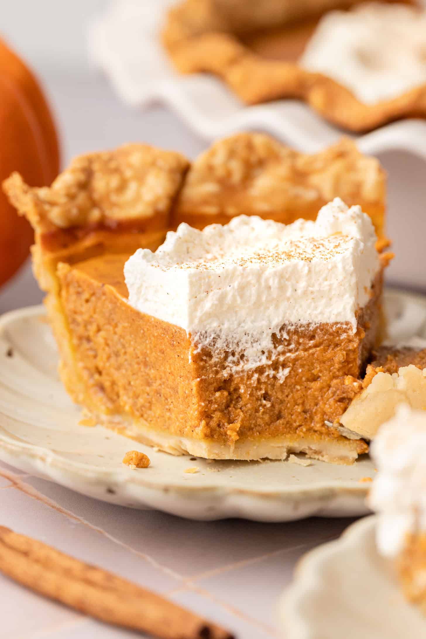 A slice of dairy free pumpkin pie on a white scalloped plate showing the thick orange filling and layers of flaky crust, with the pie dish and a small pumpkin visible in the soft-focus background
