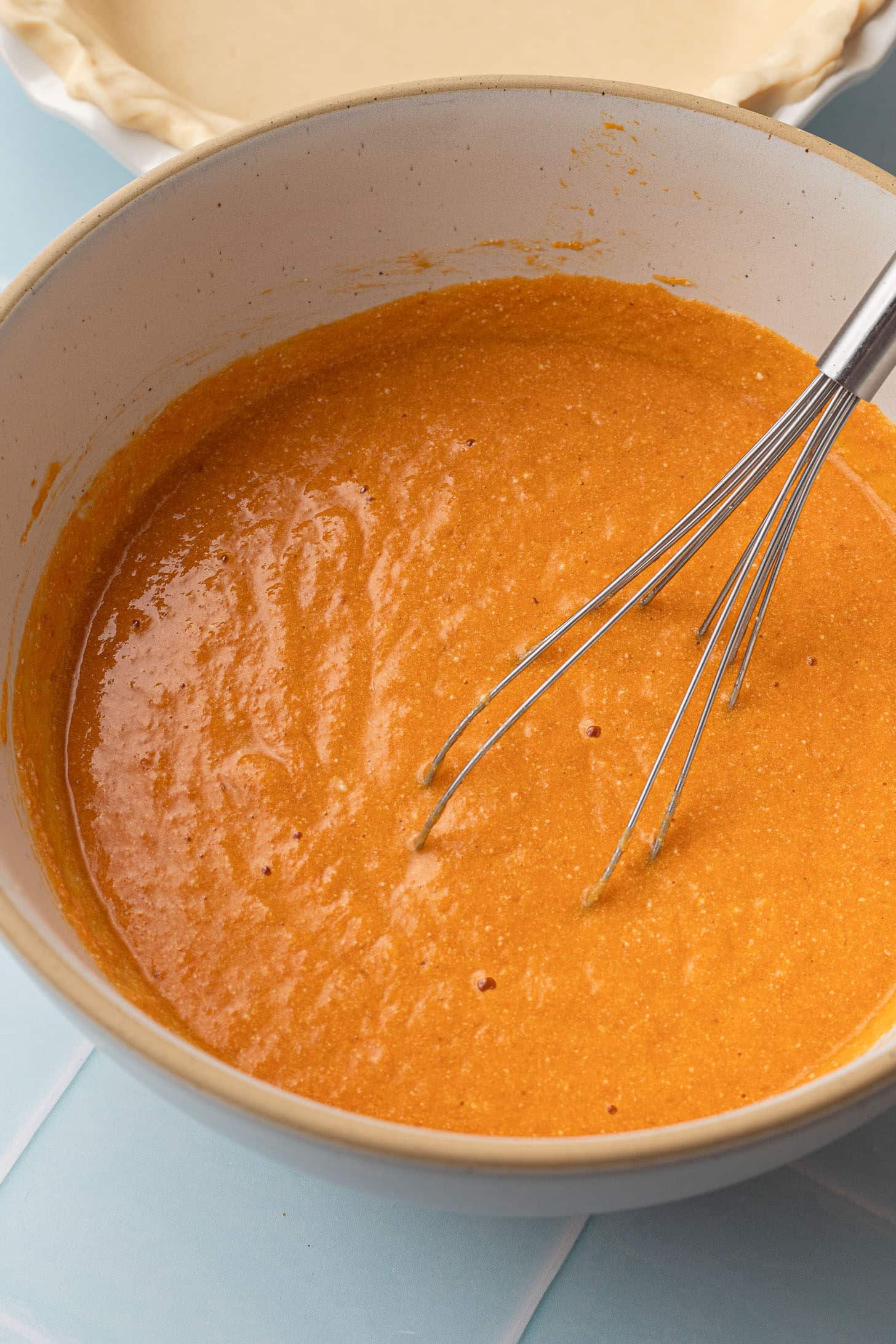 Pumpkin puree, eggs, and coconut milk being whisked together in a large cream-colored mixing bowl, with an unbaked pie crust visible in the background