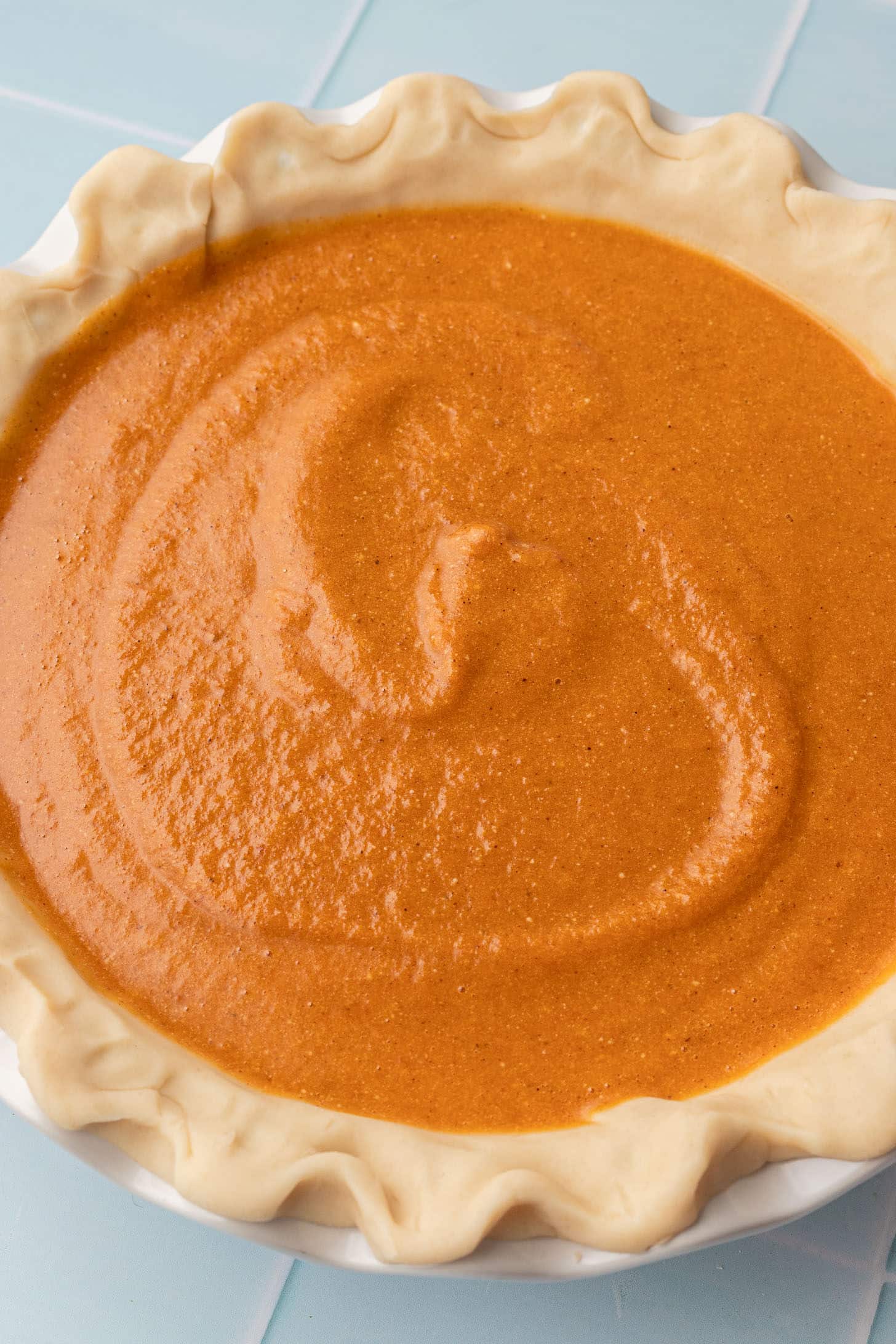 Orange pumpkin pie filling being poured into an unbaked store-bought pie crust in a white pie dish