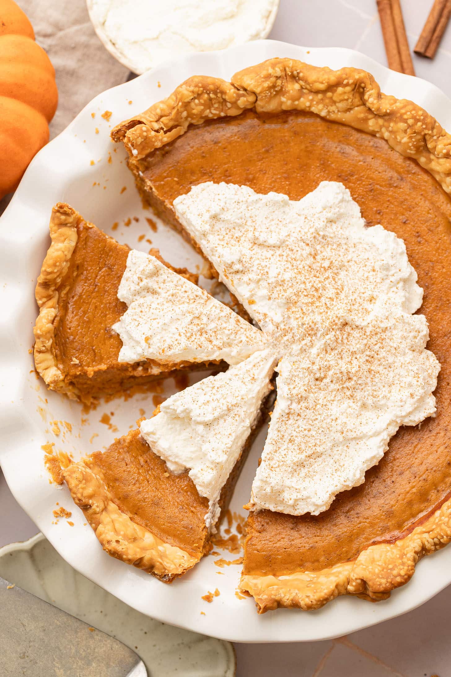 Overhead view of a baked dairy free pumpkin pie sliced into wedges and topped with dollops of whipped cream dusted with cinnamon, surrounded by mini pumpkins and cinnamon sticks on a light surface