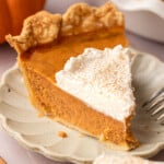 A slice of dairy free pumpkin pie on a white scalloped plate topped with a swirl of whipped cream dusted with cinnamon, with a fork resting alongside