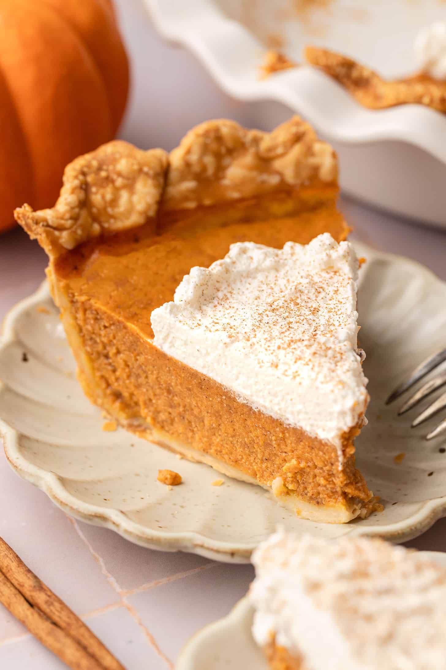 A slice of dairy free pumpkin pie on a white scalloped plate topped with a swirl of whipped cream dusted with cinnamon, with a fork resting alongside
